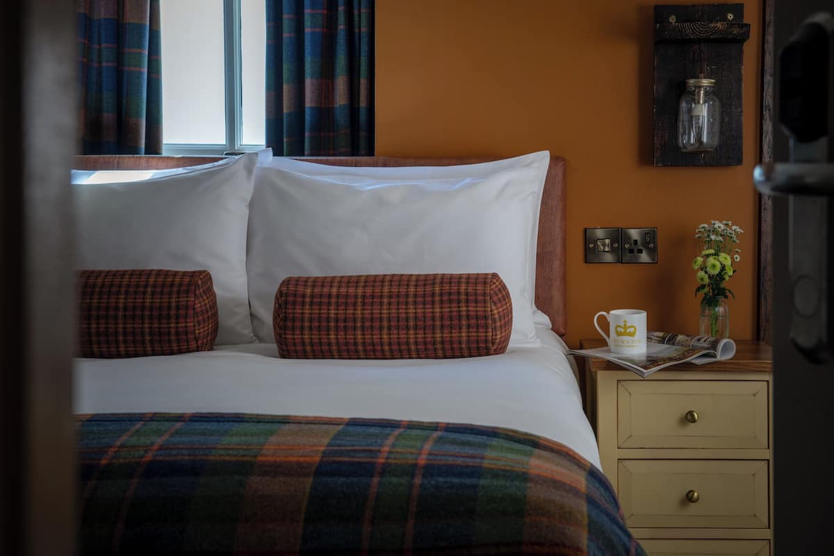 A hotel room with a bed, plaid bedding, a nightstand with a mug, a vase with flowers, and a wall-mounted light fixture.