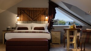 A bedroom with a large bed, a wooden headboard, a nightstand, a table with chairs, and a window with a view of trees and mountains. - Crown Inn at Pooley Bridge (Penrith)