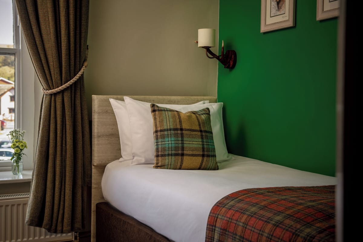 A neatly made bed with white and plaid bedding, a green wall, and a window with curtains.