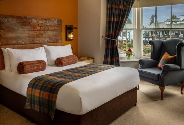 A hotel room with a large bed, a wooden headboard, a view of the outside, and a chair with a plaid cushion. - Crown Inn at Pooley Bridge (Penrith)