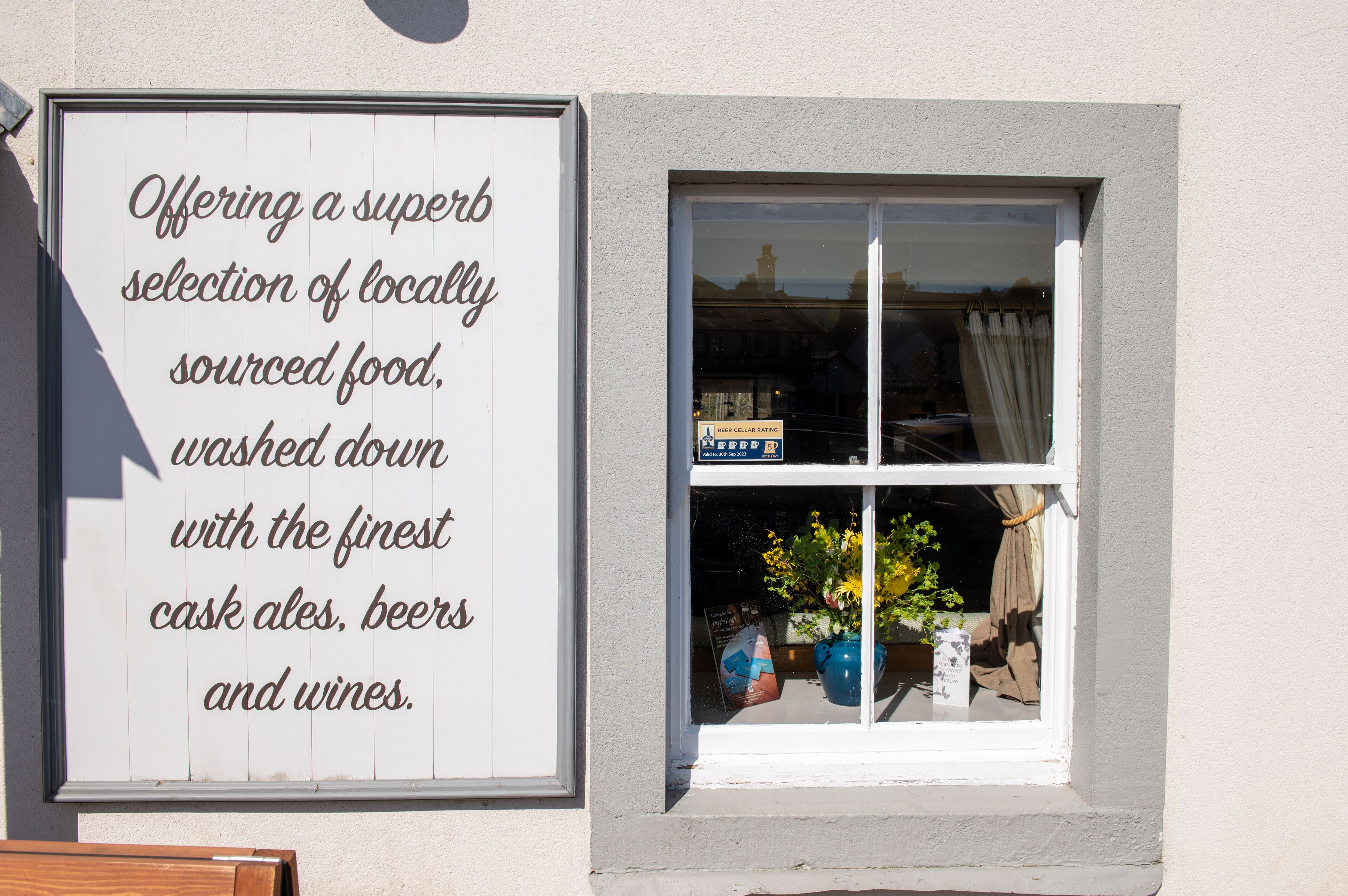 A signboard advertising locally sourced food and beverages, with a window displaying a beer cellar rating sign and a potted plant.