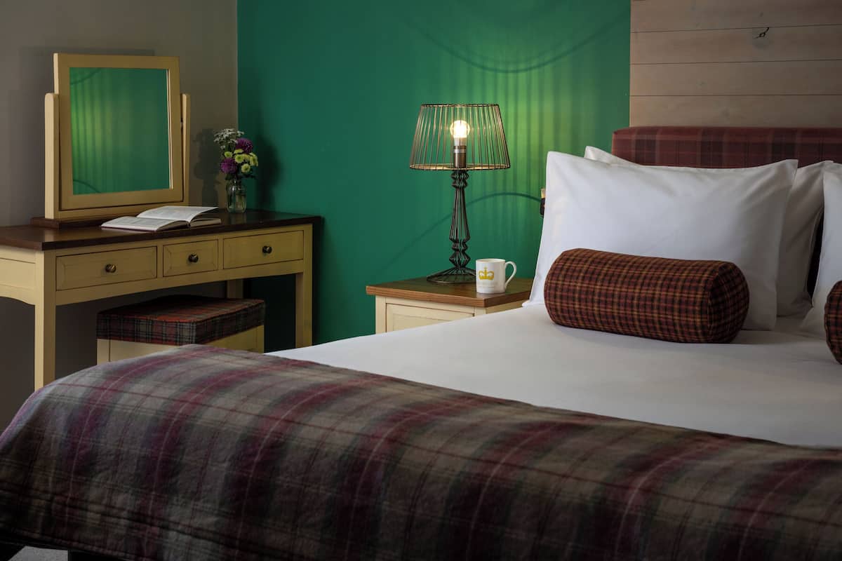 A neatly made bed with plaid bedding, a bedside table with a lamp, a small vase with flowers, and a mirror on a dresser.