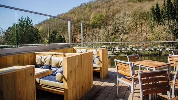 A wooden deck with seating and tables overlooking a body of water and a forested hillside. - Crown Inn at Pooley Bridge (Penrith)