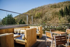 Une terrasse en bois, dotée de sièges et de tables, qui offre une vue sur une étendue d’eau et une colline boisée.