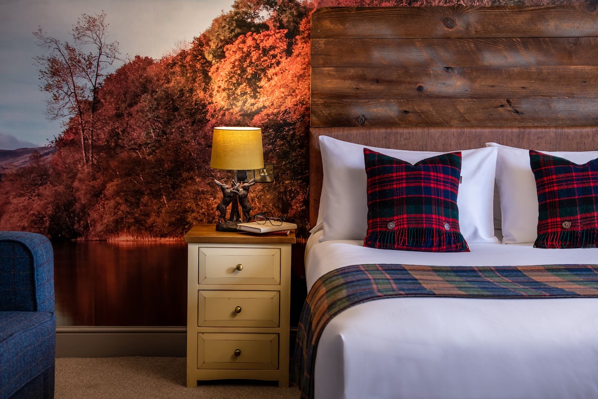 A bedroom with a wooden headboard, a bed with plaid bedding, a nightstand with a lamp, and a view of autumn trees.