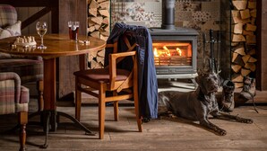 A cozy room with a wooden table, chairs, a glass of beer, a dog, and a wood-burning stove. - Crown Inn at Pooley Bridge (Penrith)