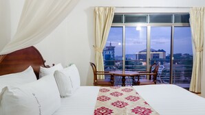 Habitación de hotel con una cama grande, una mesita, dos sillas y vistas de la ciudad a través de amplios ventanales.