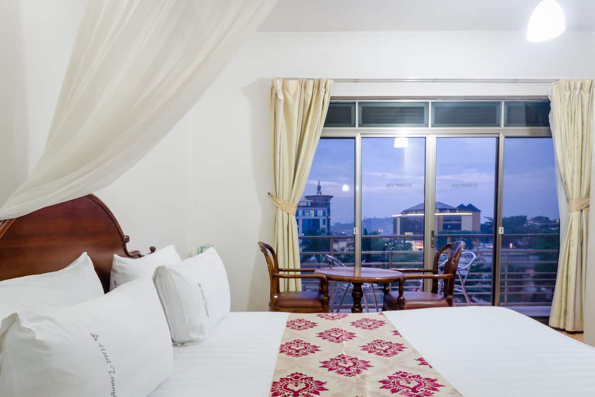 A hotel room with a large bed, a small table, two chairs, and a view of the city through large windows.