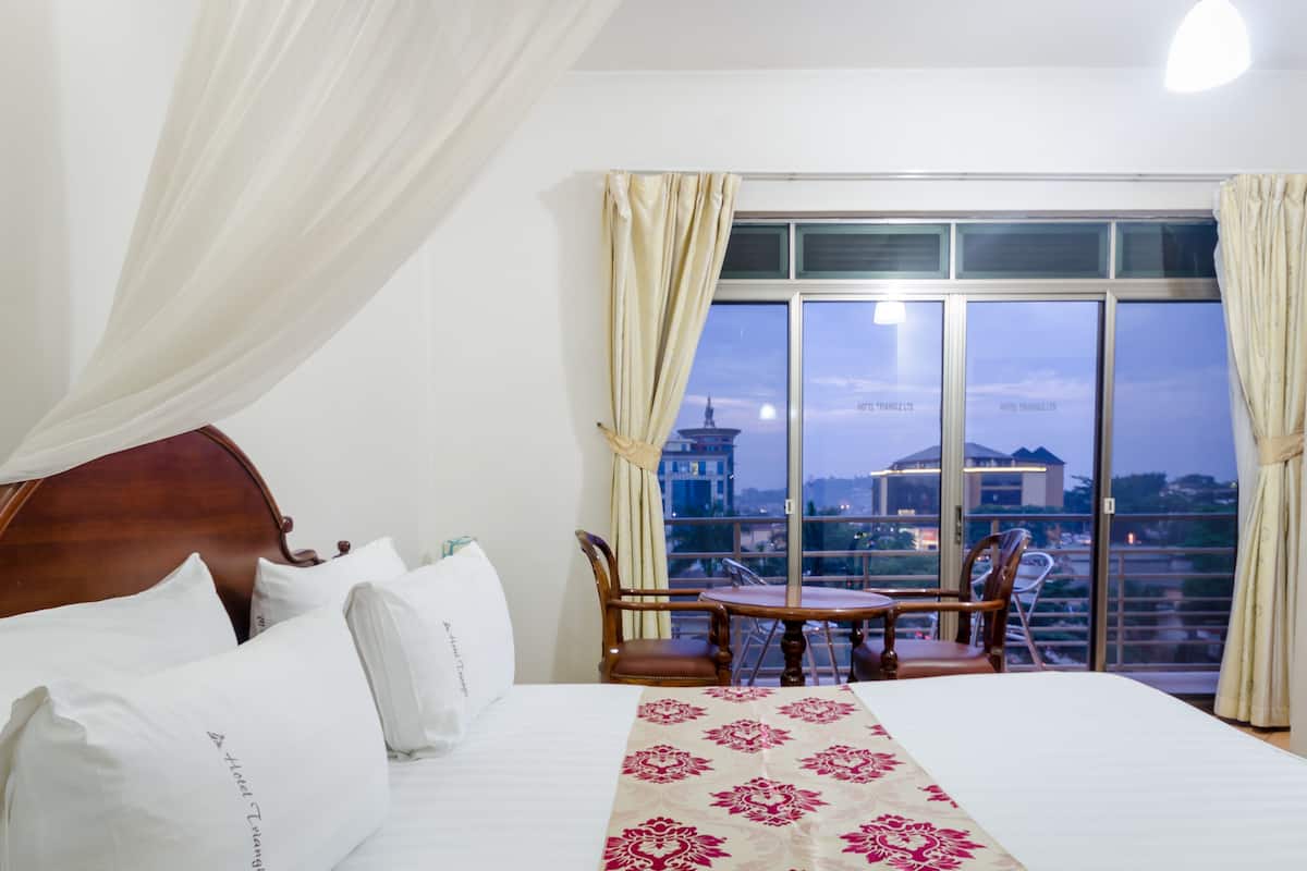 A hotel room with a large bed, a dining table with chairs, and a view of the cityscape through large windows.