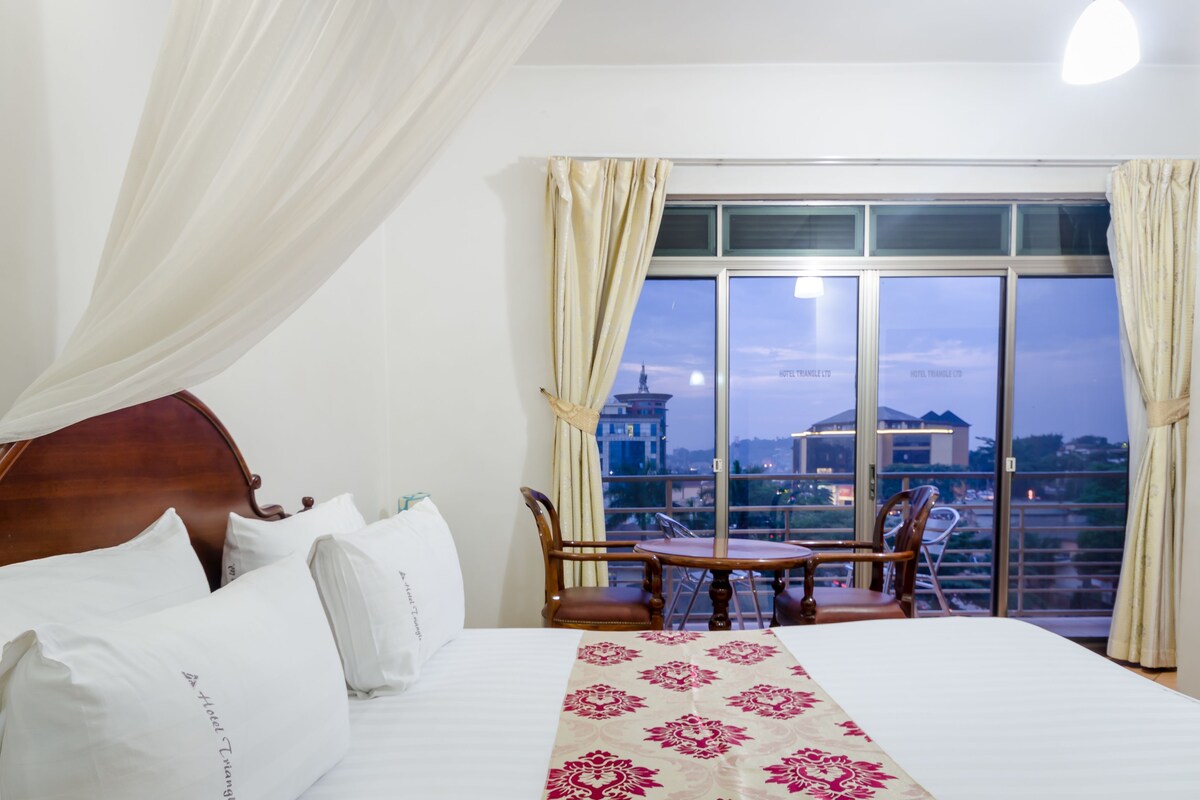 Habitación de hotel con una cama grande, una mesa de comedor con sillas y vistas a la ciudad a través de amplios ventanales.
