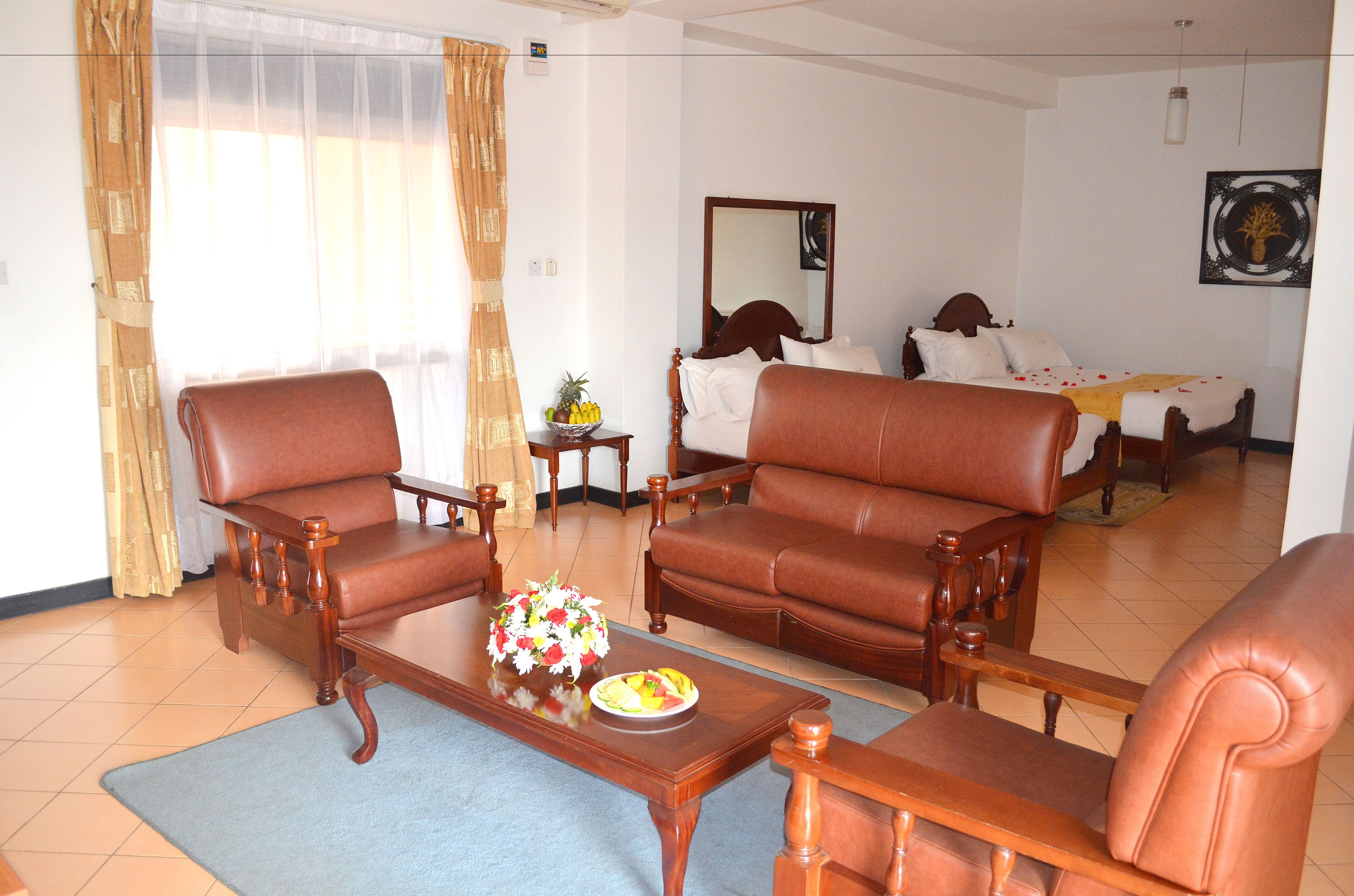 A living room with two brown leather sofas, a wooden coffee table with a fruit bowl, and a bed with white bedding in the background.