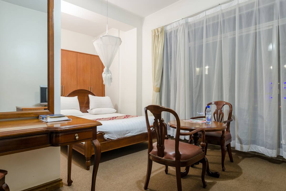 A hotel room with a wooden bed, a desk, two chairs, and a large window with sheer curtains.