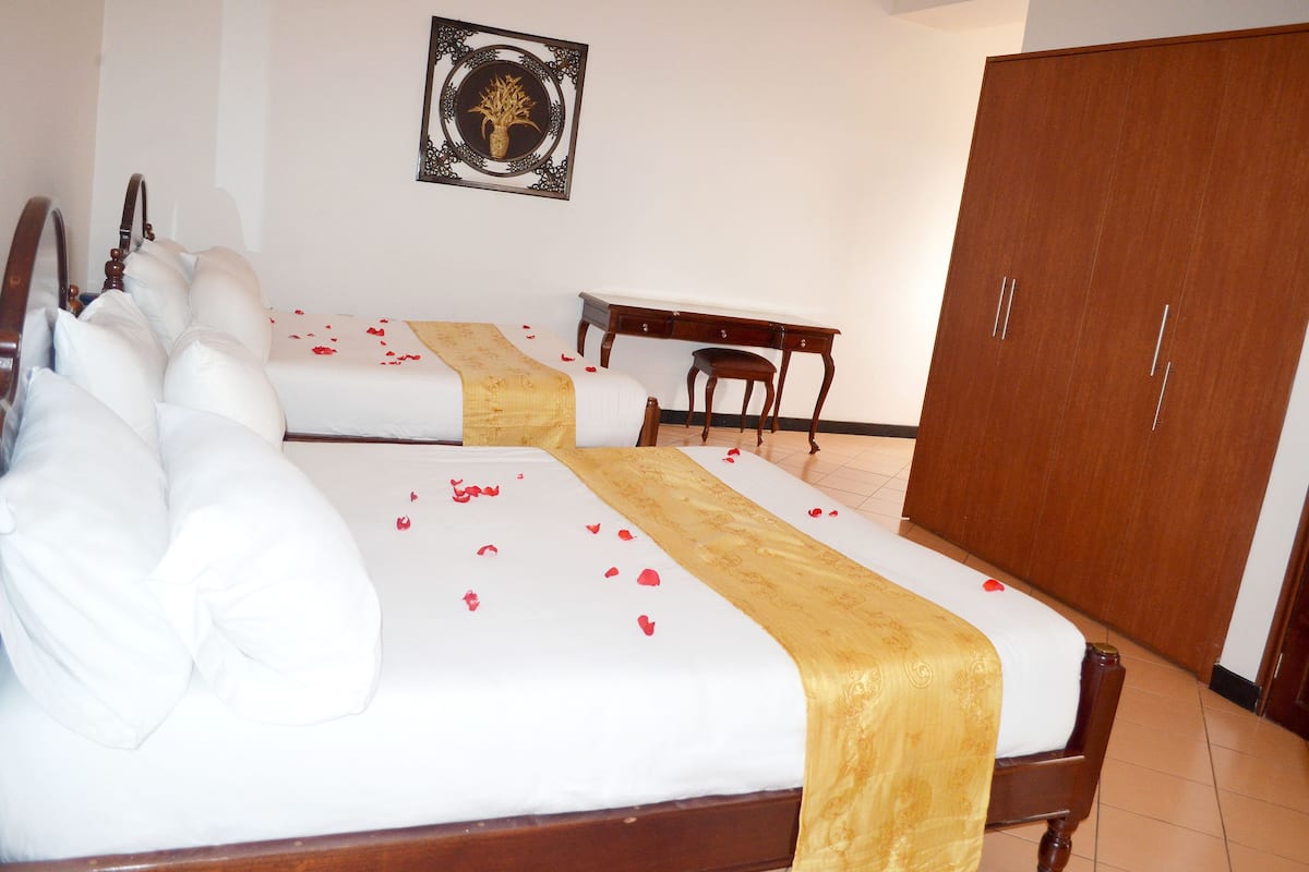 A hotel room with two beds, a wooden wardrobe, a desk, and a framed artwork on the wall.