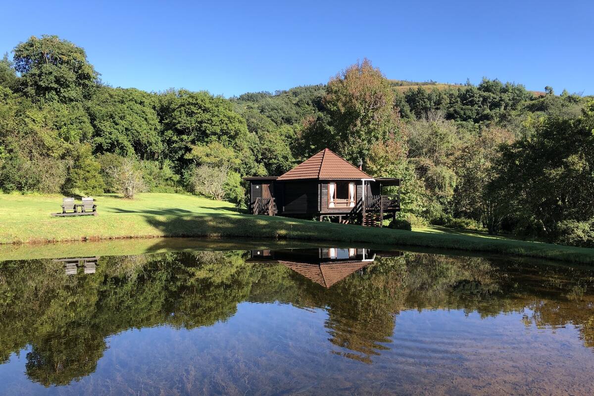 A wooden cabin with a red roof, situated by a calm pond reflecting the surrounding trees.