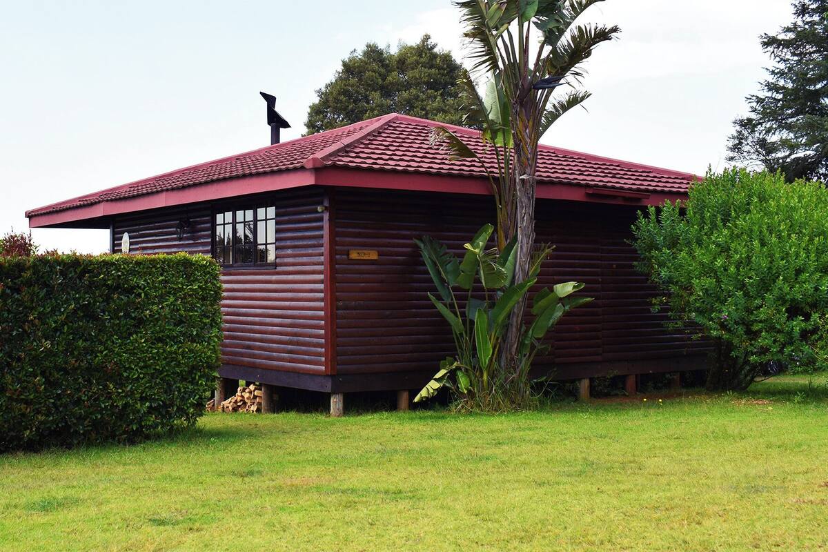 A rustic cabin with a red-tiled roof, surrounded by lush greenery and a well-maintained lawn.