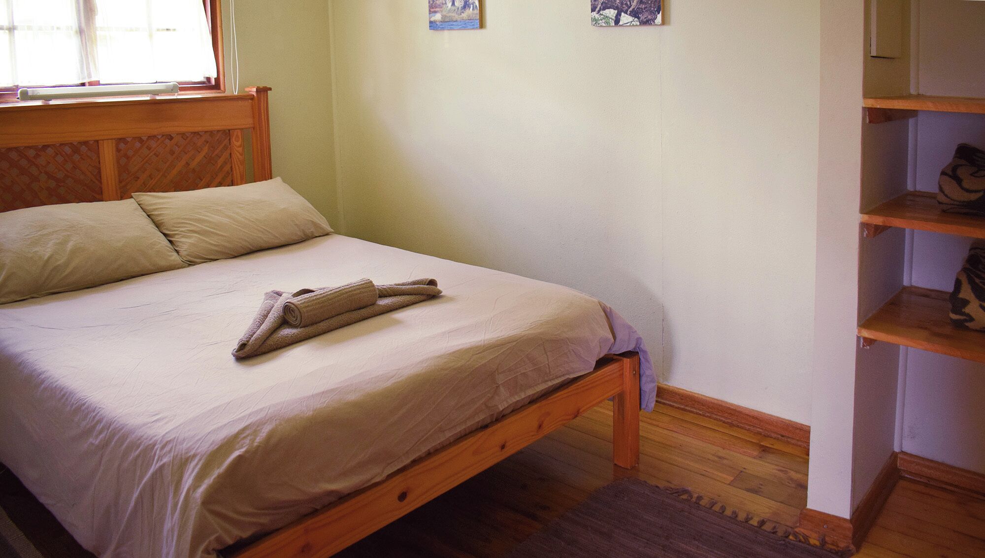 A neatly made bed with a wooden headboard, a rolled towel, and a plain wall with a small picture.