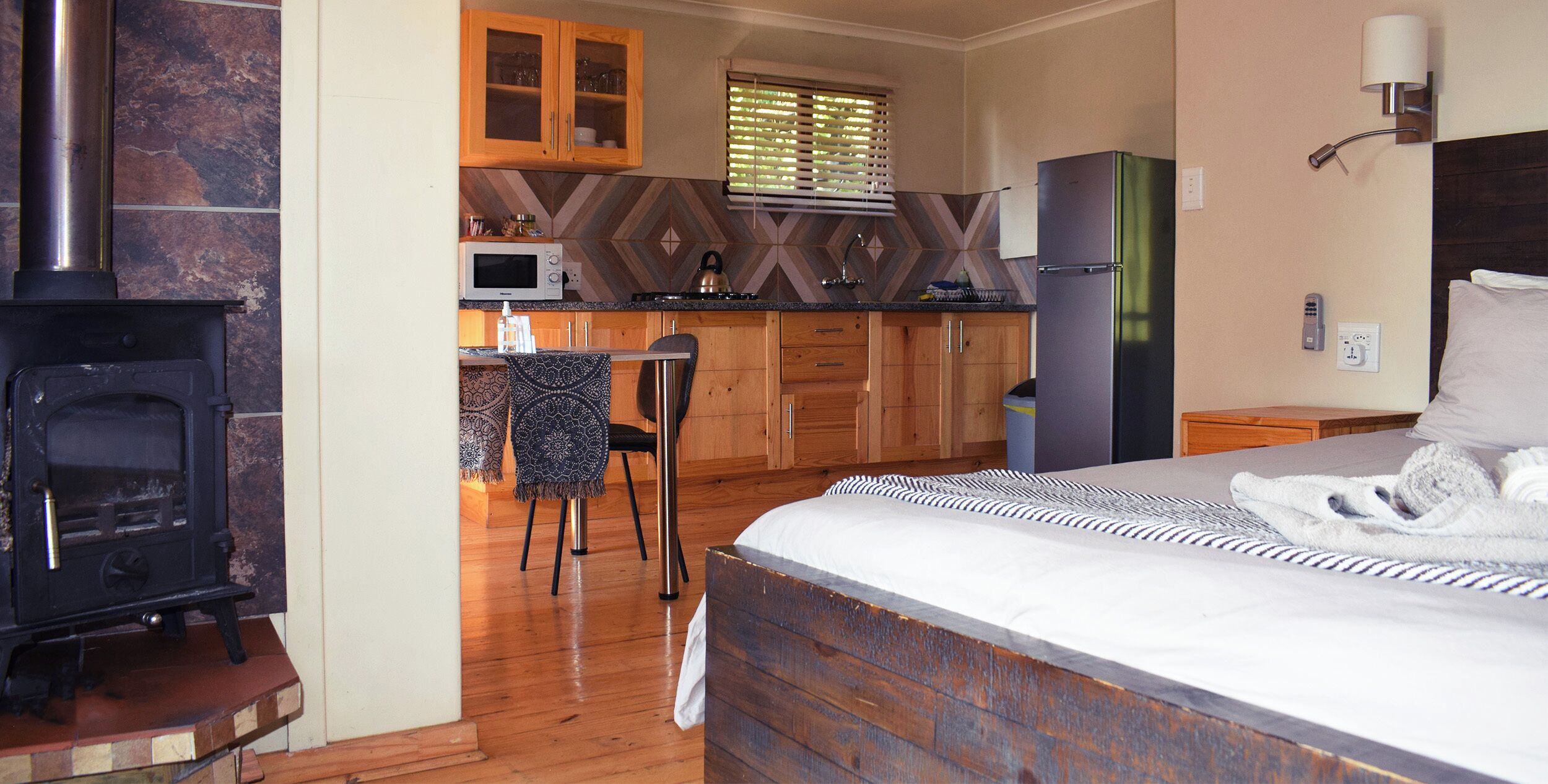 A bedroom with a large bed, a wooden headboard, and a nightstand. Adjacent to the bedroom is a kitchen with wooden cabinets, a microwave, and a refrigerator.
