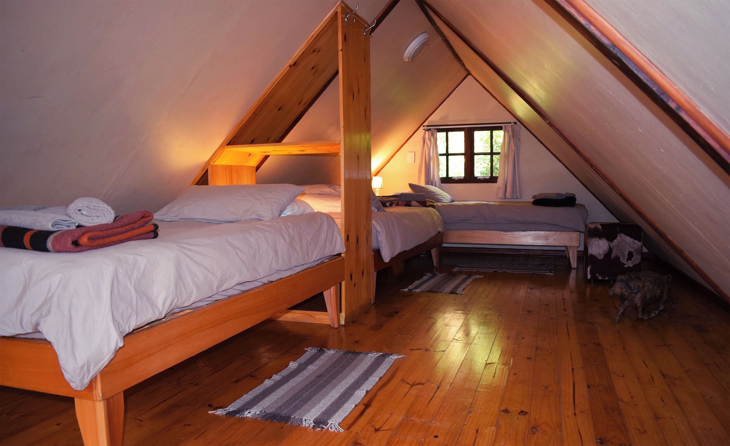 A small attic bedroom with two wooden bunk beds, a window with curtains, and a wooden floor.