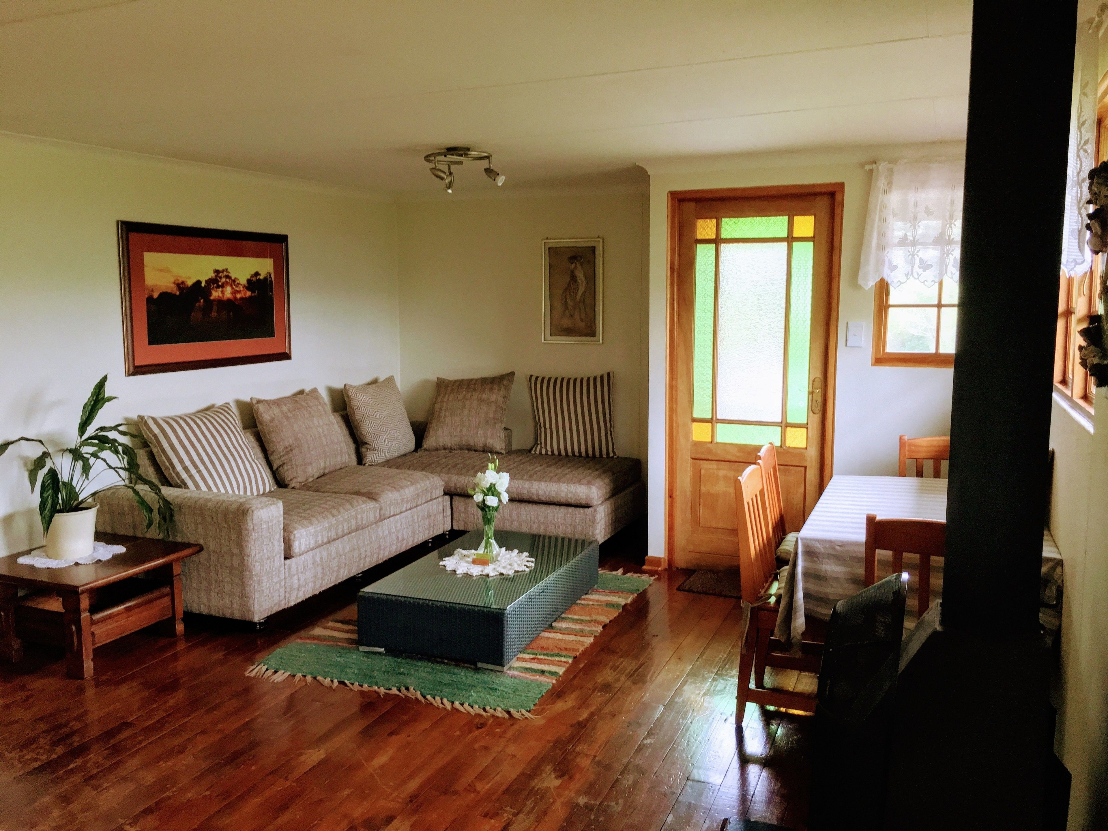 Una sala de estar acogedora con un sofá, una mesa de centro, una mesa de comedor y una silla. Se ve vegetación a través de la ventana.