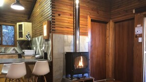 L’intérieur chaleureux d’un chalet comprend un poêle à bois, un coin repas avec deux chaises et une cuisine dotée de meubles en bois et d’une fenêtre.