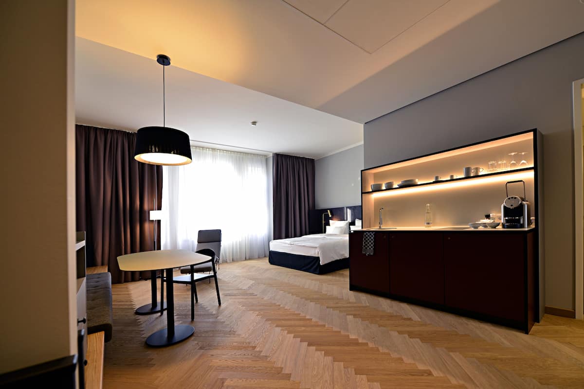 A modern hotel room with a bed, a desk with a chair, a dining table, and a kitchen area with a coffee machine.