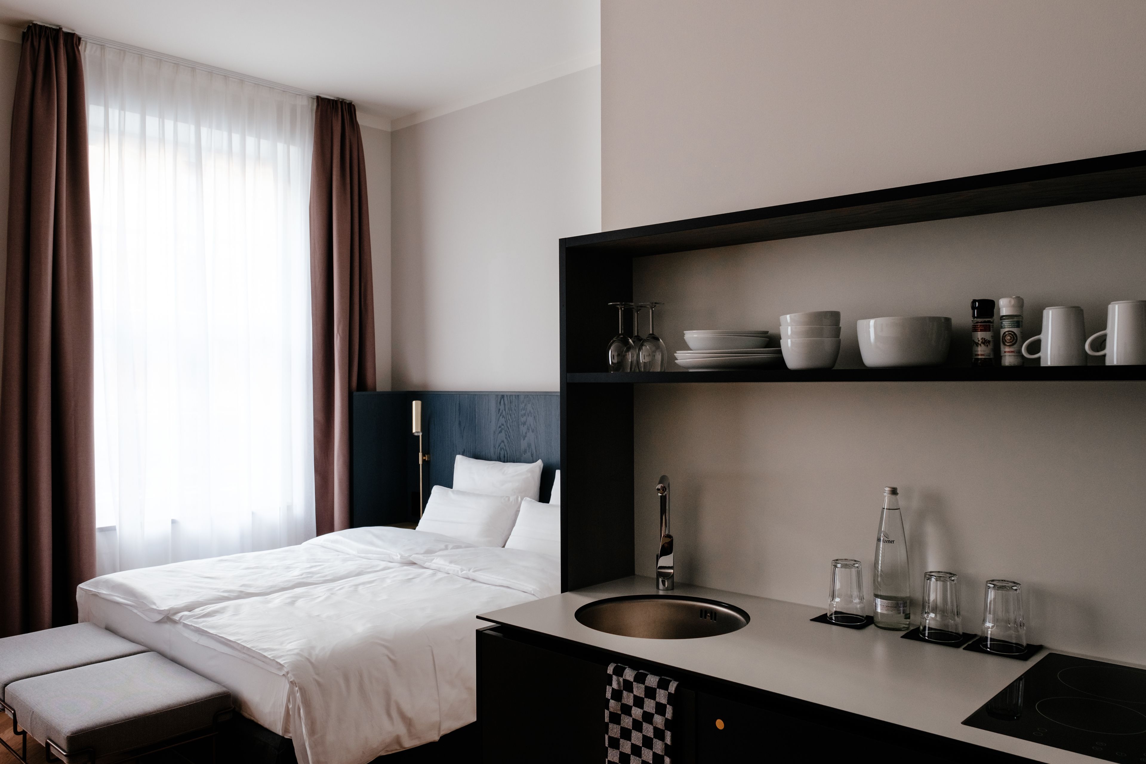 Una habitación de hotel con una cama, una mesita de noche con lavamanos y un estante con vasos y tazas.