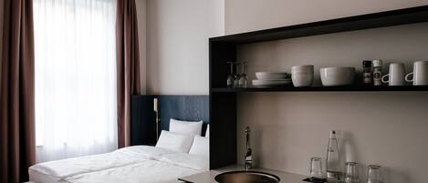 Una habitación de hotel con una cama, una mesita de noche con lavamanos y un estante con vasos y tazas.