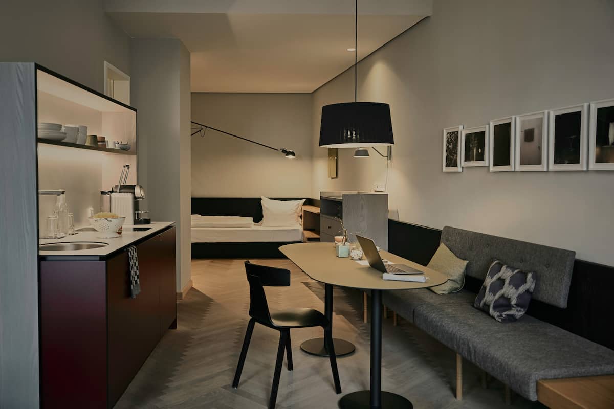 A modern hotel room with a kitchenette, a dining area with a table and chairs, a sofa, and a bed.