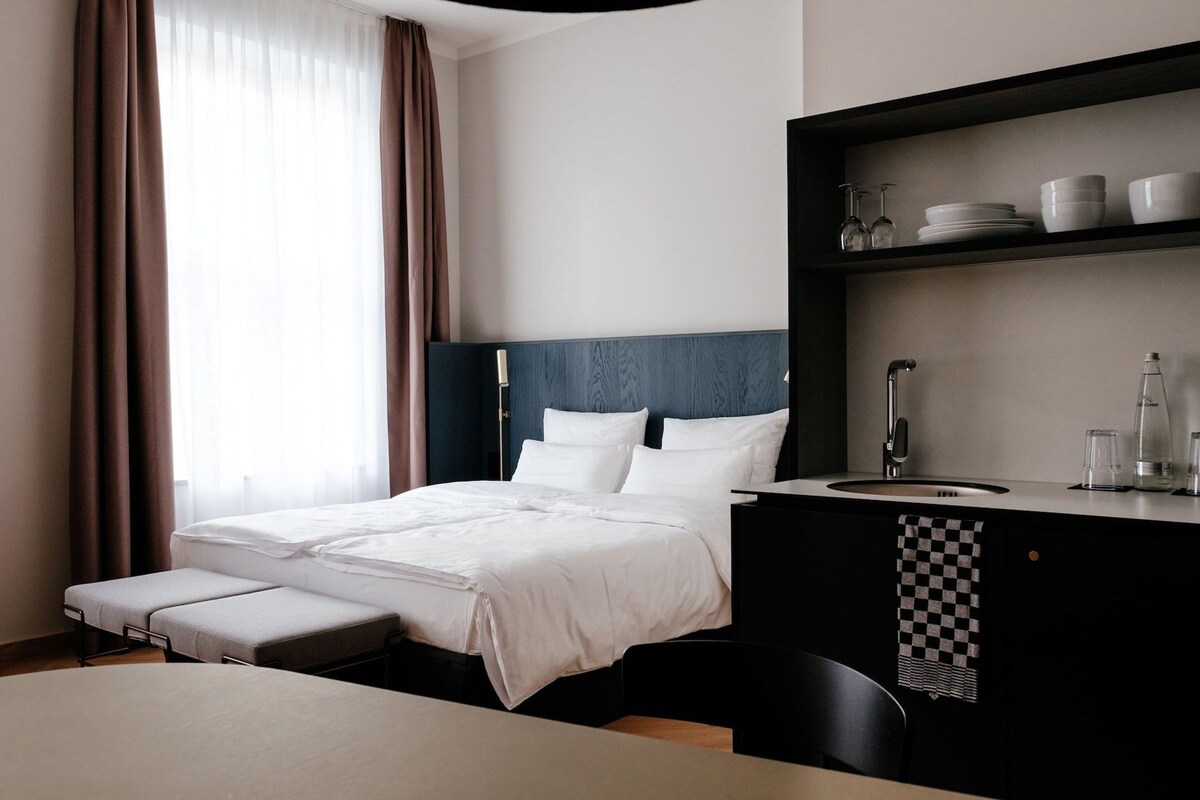 A hotel room with a bed, bedside table, and a small kitchenette area with a sink and storage.