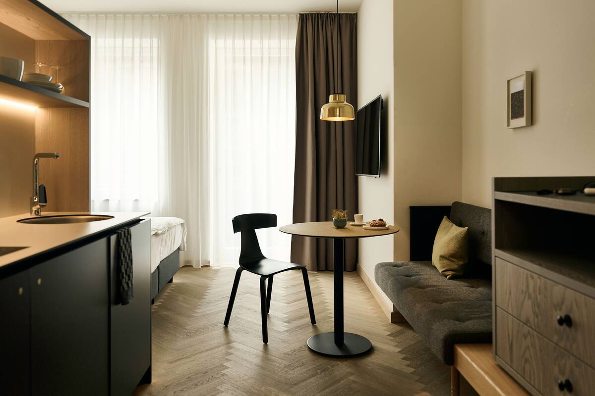 A modern, compact hotel room with a kitchenette, a dining area with a round table and two chairs, a sofa, and a TV mounted on the wall.