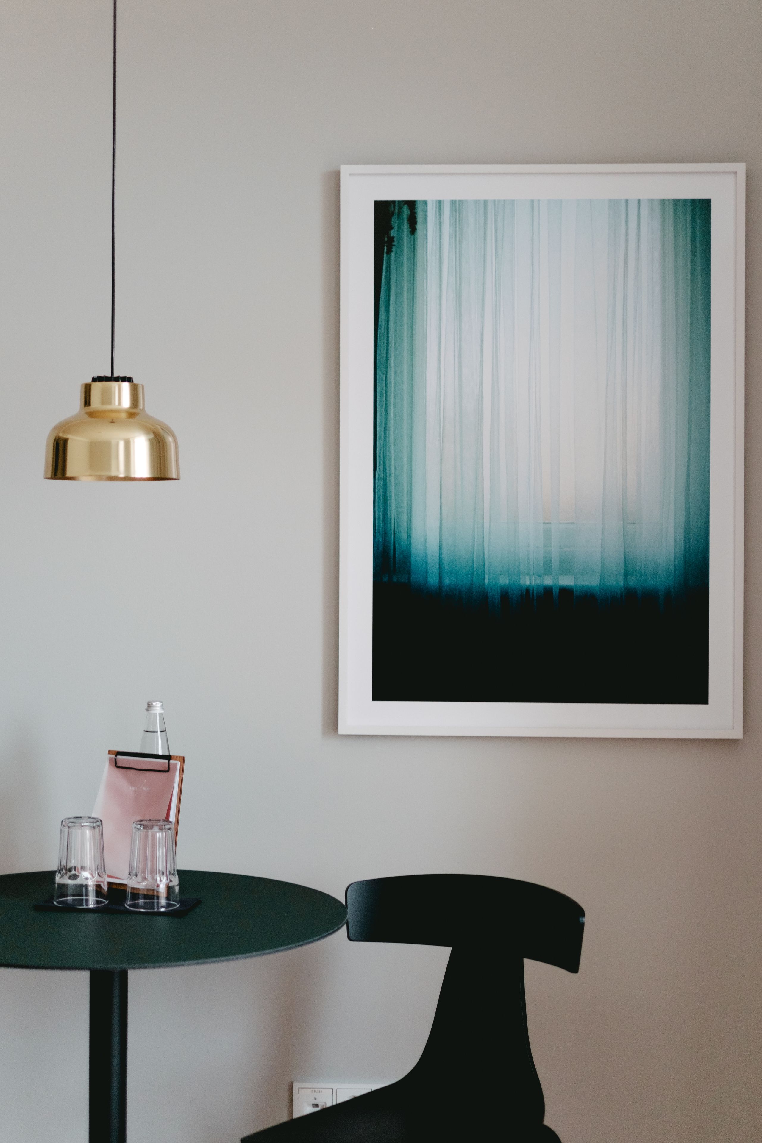 Una habitación con una mesa redonda, una silla negra, un cuadro abstracto enmarcado y una lámpara colgante dorada.
