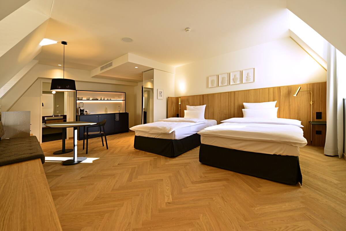 A modern hotel room with two beds, a desk, and a dining area.