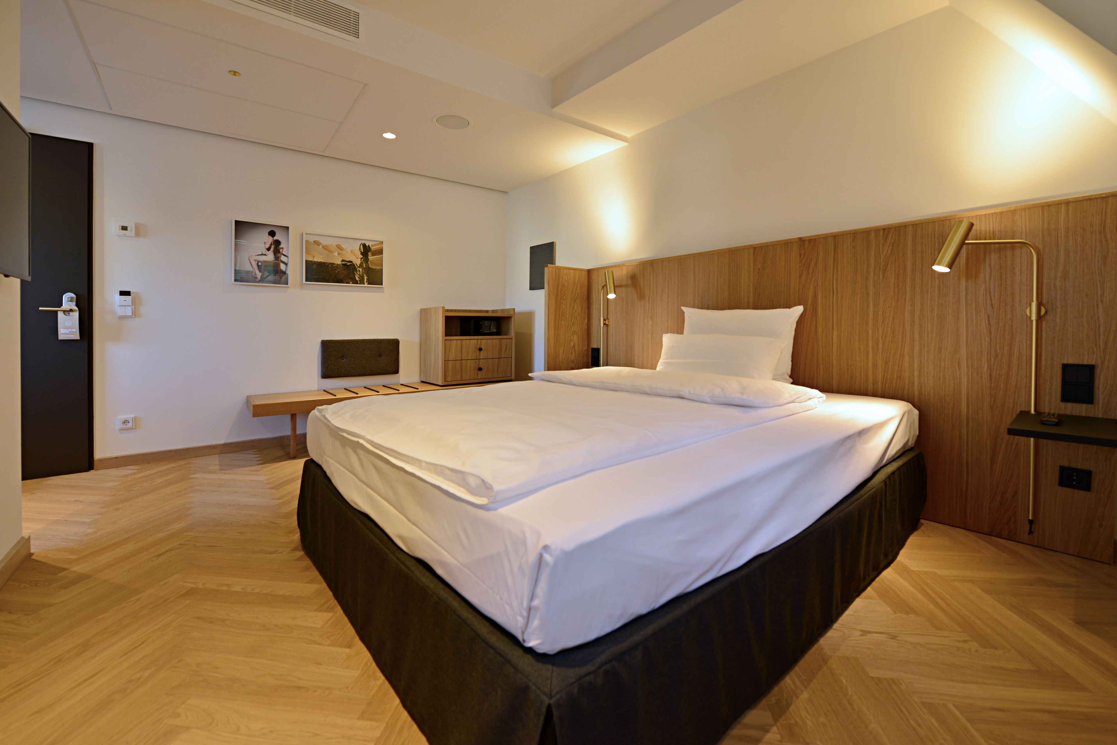 A modern hotel room with a large bed, wooden headboard, bedside tables, a desk, and a wall-mounted lamp.
