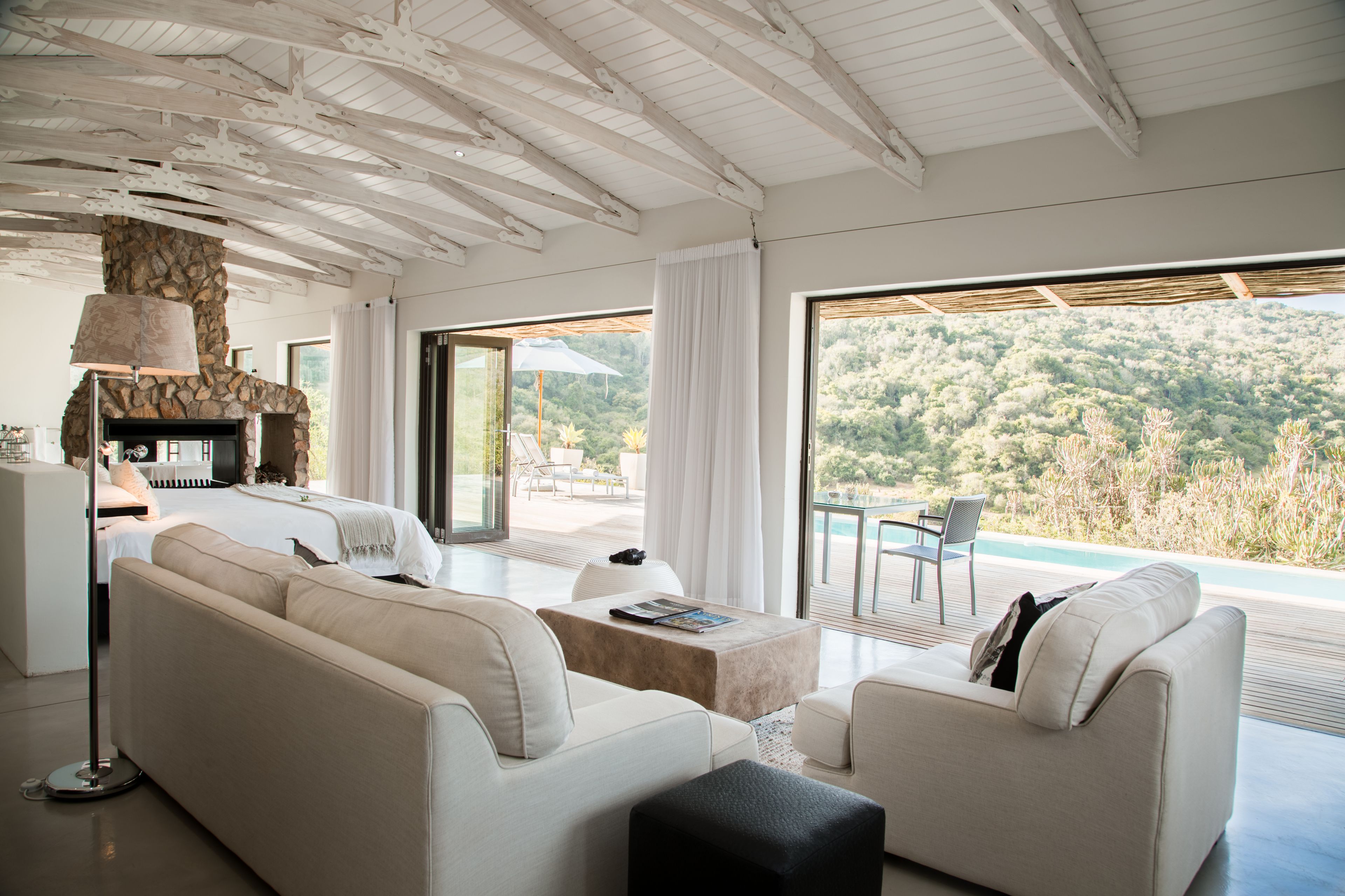 A modern, well-lit living room with a stone fireplace, a large sofa, a coffee table, and a view of a lush landscape through large windows.