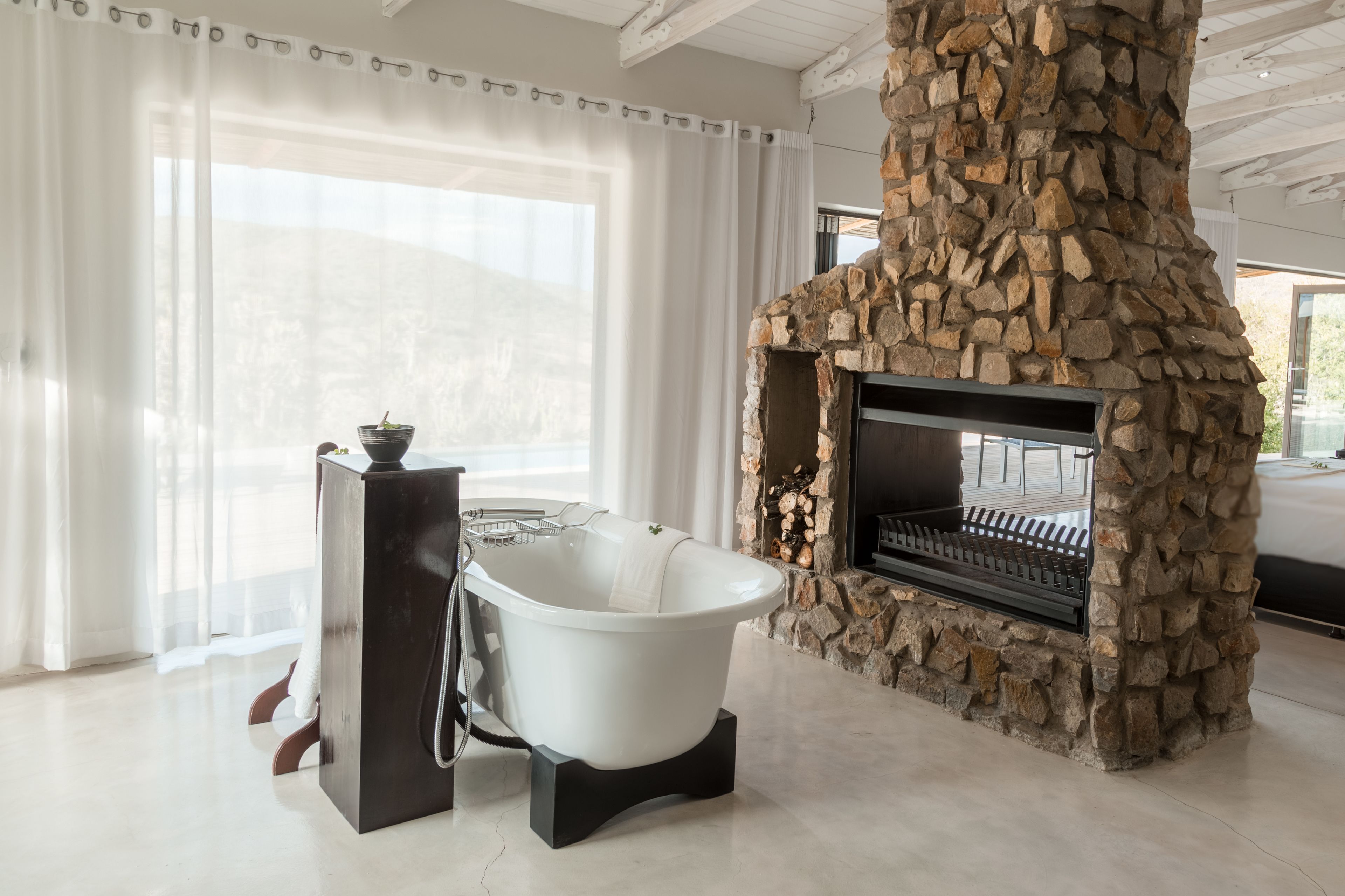 A bathroom with a freestanding bathtub, a stone fireplace, and a large window with sheer curtains.