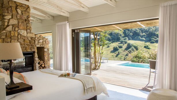 Une chambre avec une cheminée en pierre, un grand lit et une vue sur une piscine et la verdure environnante.