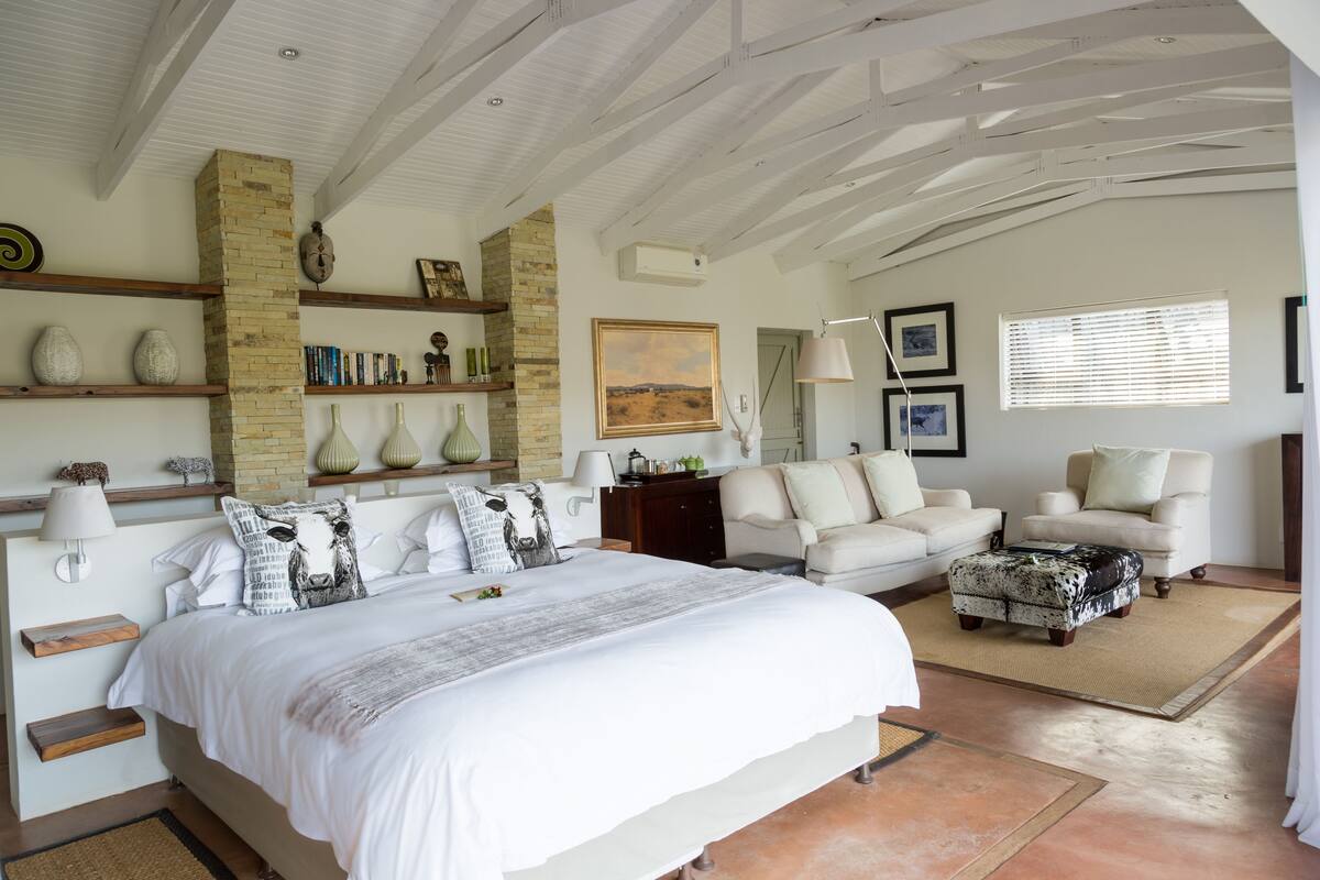 A spacious bedroom with a large bed, two white armchairs, a sofa, a fireplace, and a bookshelf.