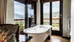 Une salle de bain avec une baignoire sur pieds, une chaise en bois, un tableau et une vue sur un paysage montagneux, visible à travers de grandes fenêtres.