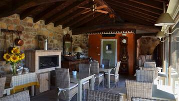Un área de comedor al aire libre con mobiliario de mimbre, pared de piedra, chimenea y ventilador de techo.
