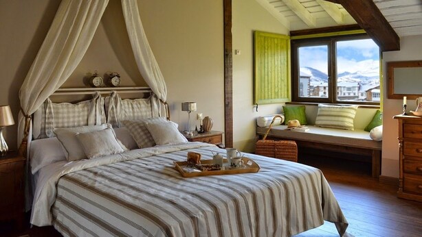 Un dormitorio con cama con dosel, mesita de noche, lámpara, ventana con vistas y un banco con cojines.