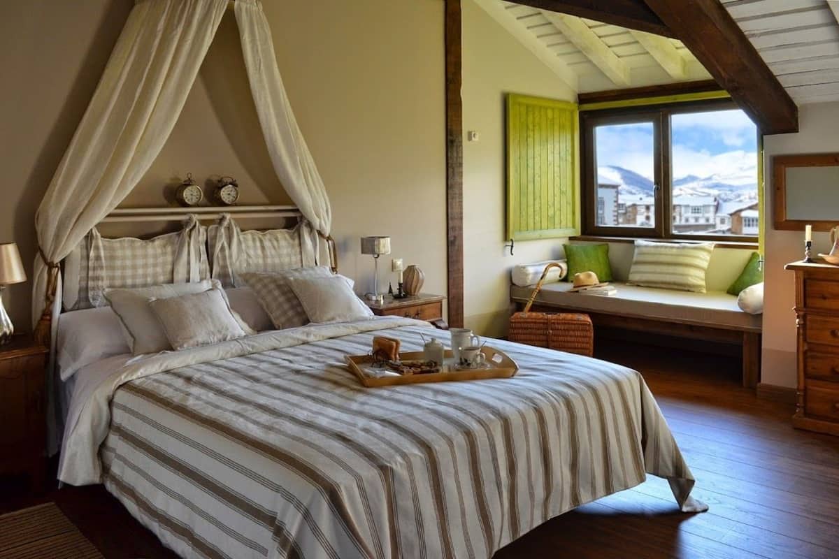 A bedroom with a canopy bed, bedside table, lamp, window with view, and a bench with cushions.
