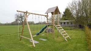 A wooden swing set with a slide and a small shelter in a grassy field.