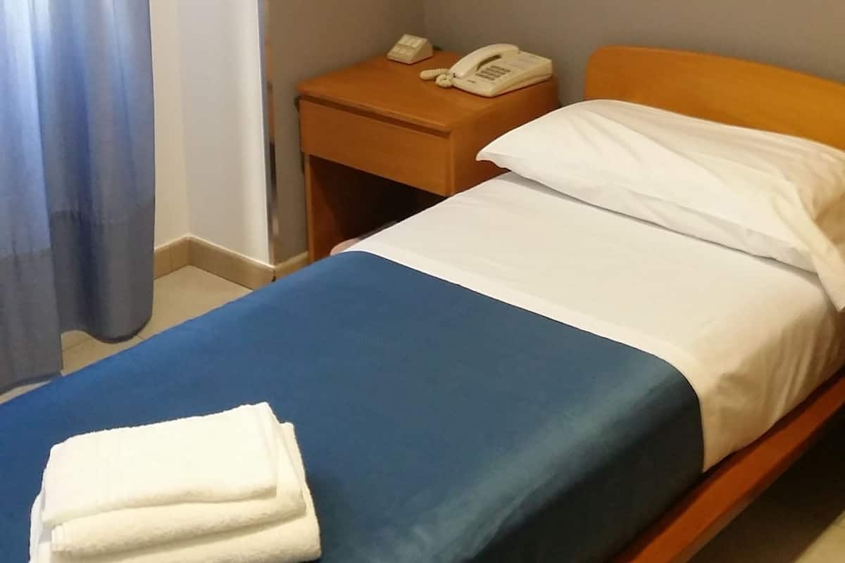 A single bed with a blue bedspread and white pillows, a wooden nightstand with a telephone, and a blue curtain partially drawn to the side.