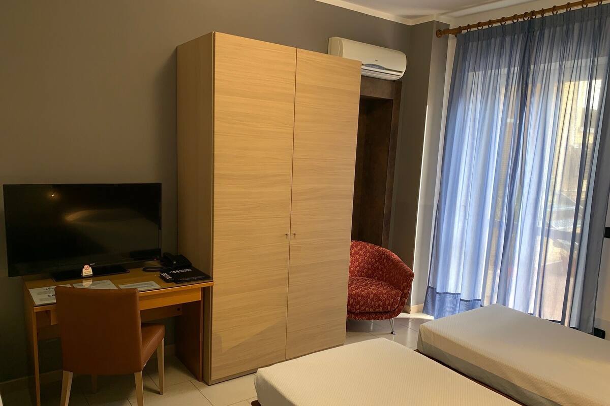 A hotel room with a bed, a desk with a computer, a TV, a wardrobe, and a red chair.