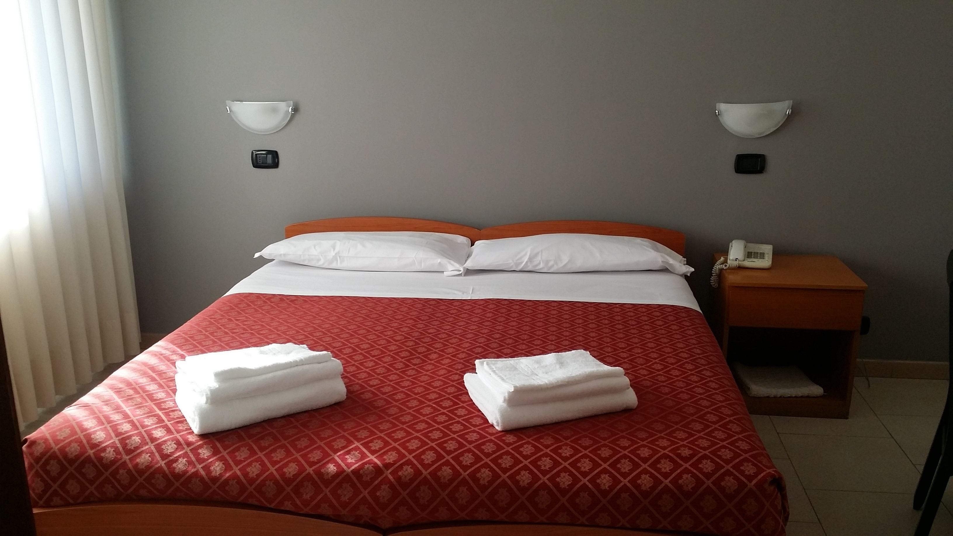 Una cama bien hecha con una colcha roja estampada, dos almohadas blancas y dos toallas dobladas. A la derecha, un mesita de noche de madera con un teléfono y una lámpara fijada a la pared.