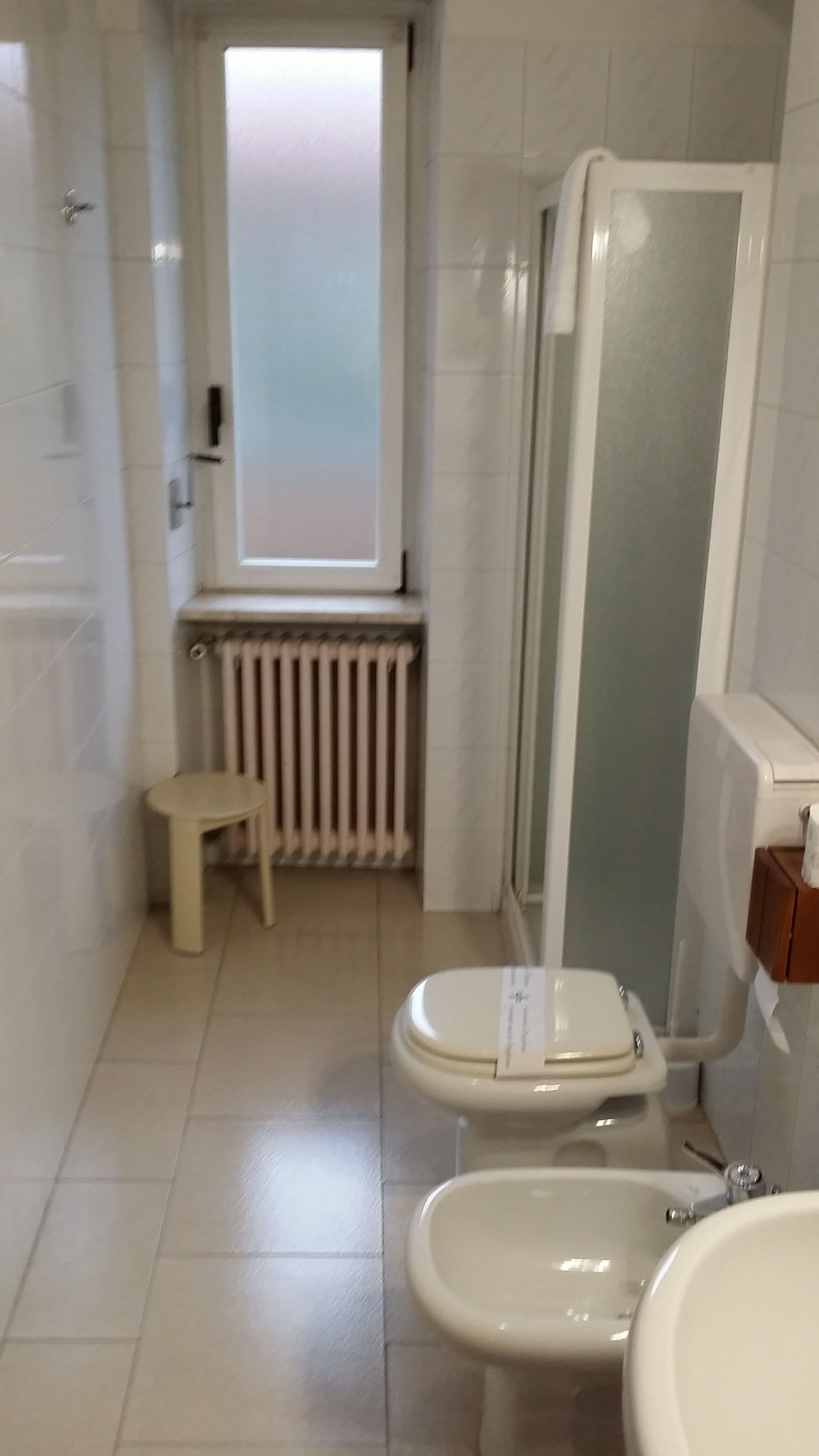 A bathroom with two toilets, a sink, a small stool, a radiator, and a frosted glass window.