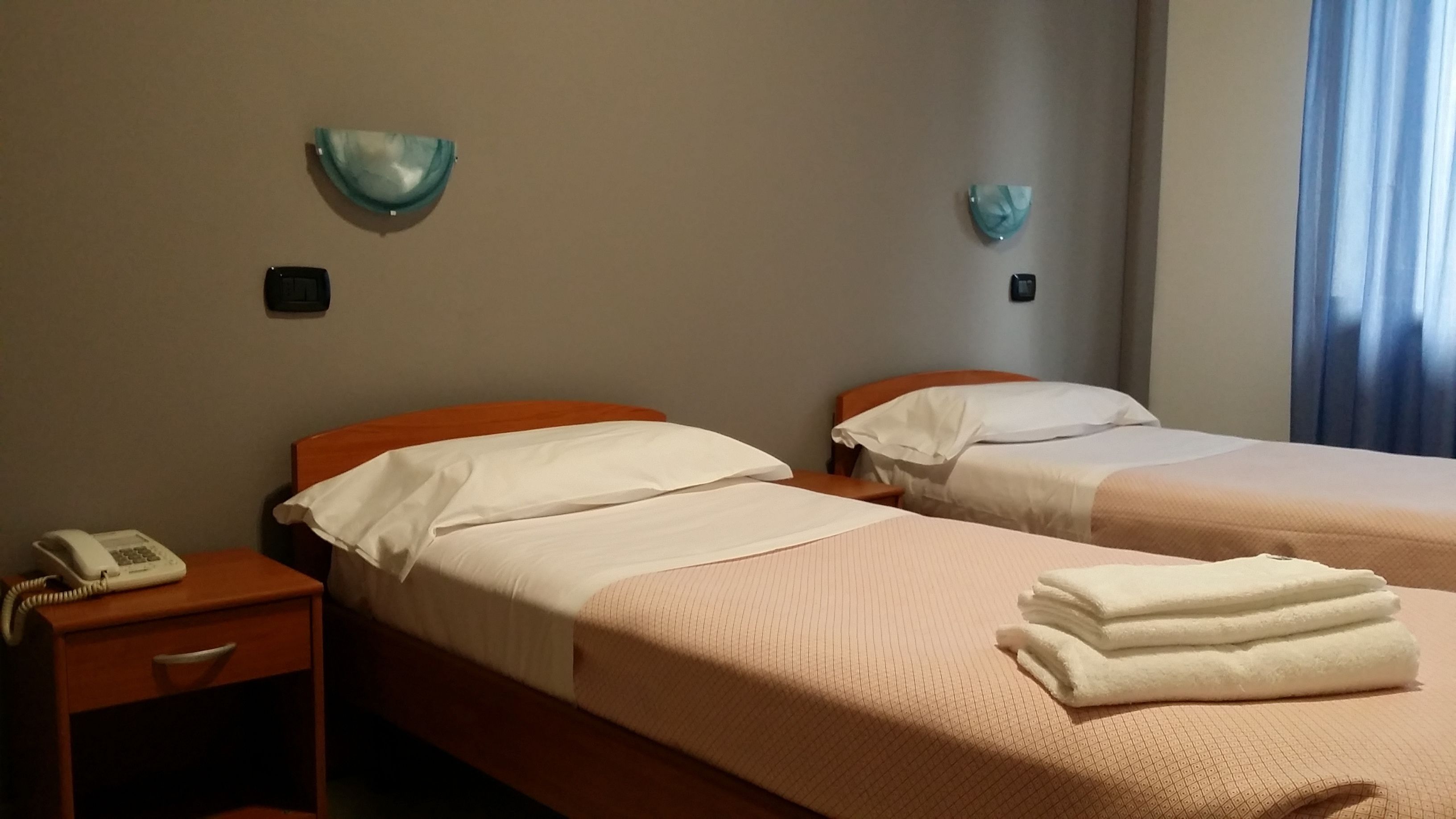 A hotel room with two beds, a bedside table with a telephone, and a wall-mounted light fixture.