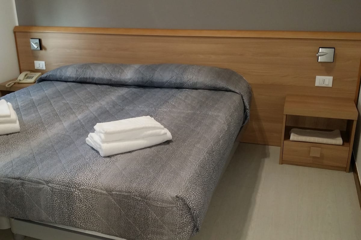 A hotel room with a bed, a wooden headboard, a bedside table with a phone, and a small drawer unit.
