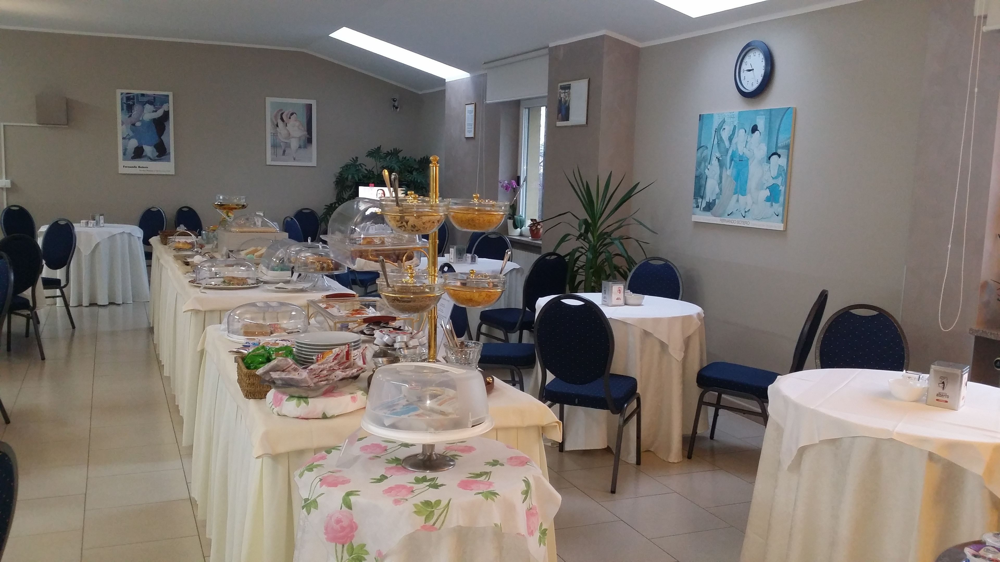 A banquet hall with round tables covered in white tablecloths, each with a floral table runner. There are chairs around the tables, and the room has a clock on the wall.
