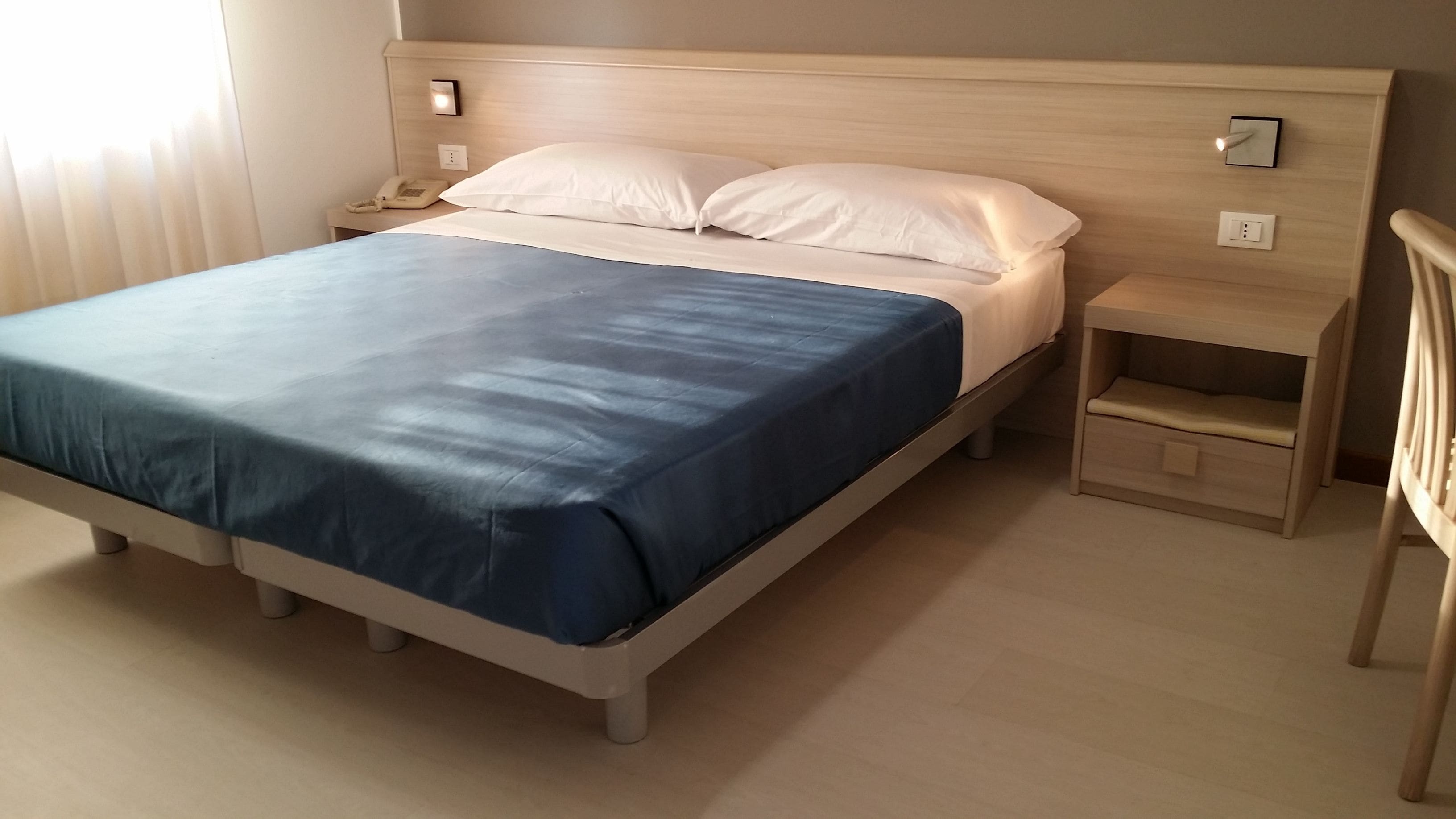 A bed with a blue bedspread, a wooden headboard, a bedside table with a lamp, and a nightstand with a phone.